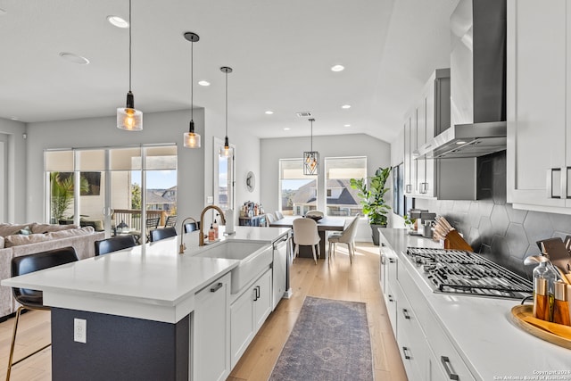 kitchen with sink, wall chimney exhaust hood, hanging light fixtures, a kitchen island with sink, and light wood-type flooring