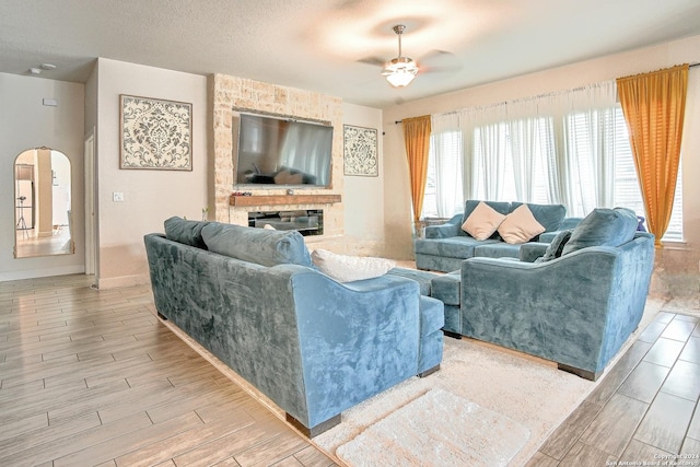 living room featuring a textured ceiling, light hardwood / wood-style flooring, ceiling fan, and a stone fireplace