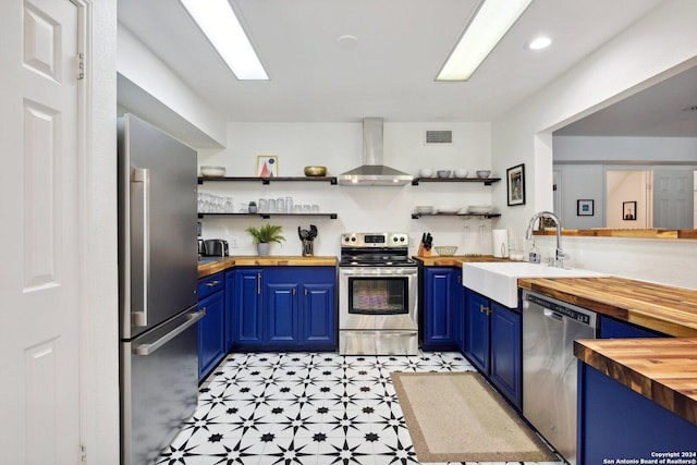 kitchen featuring wall chimney exhaust hood, stainless steel appliances, sink, blue cabinetry, and butcher block counters