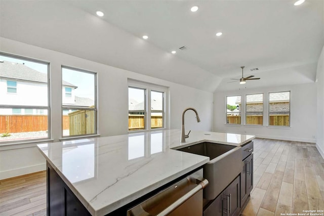 kitchen featuring stainless steel dishwasher, a wealth of natural light, light wood-type flooring, an island with sink, and light stone counters