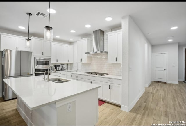 kitchen with appliances with stainless steel finishes, wall chimney exhaust hood, sink, decorative light fixtures, and light hardwood / wood-style flooring