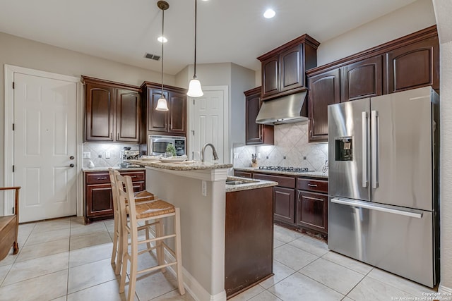 kitchen featuring appliances with stainless steel finishes, tasteful backsplash, light stone counters, a center island with sink, and hanging light fixtures