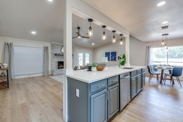 kitchen featuring stainless steel dishwasher, light wood-type flooring, a fireplace, an island with sink, and decorative light fixtures