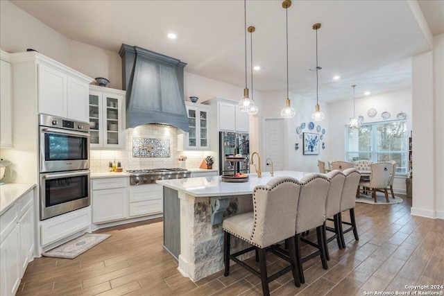 kitchen featuring appliances with stainless steel finishes, premium range hood, light hardwood / wood-style floors, hanging light fixtures, and an island with sink