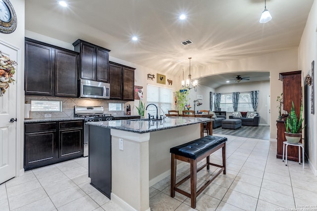 kitchen featuring hanging light fixtures, dark stone counters, a kitchen island with sink, ceiling fan with notable chandelier, and appliances with stainless steel finishes