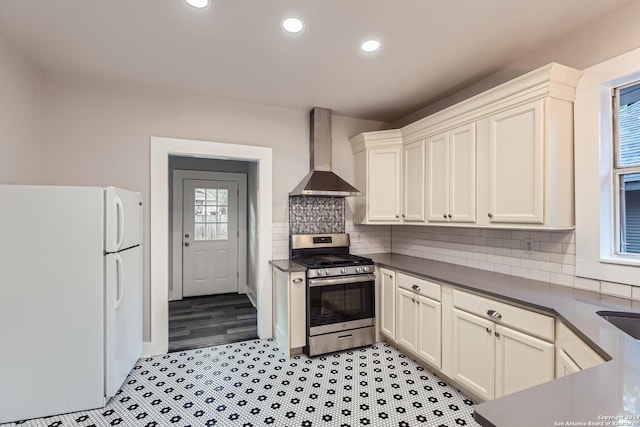 kitchen with stainless steel gas range oven, backsplash, white cabinets, white refrigerator, and wall chimney exhaust hood