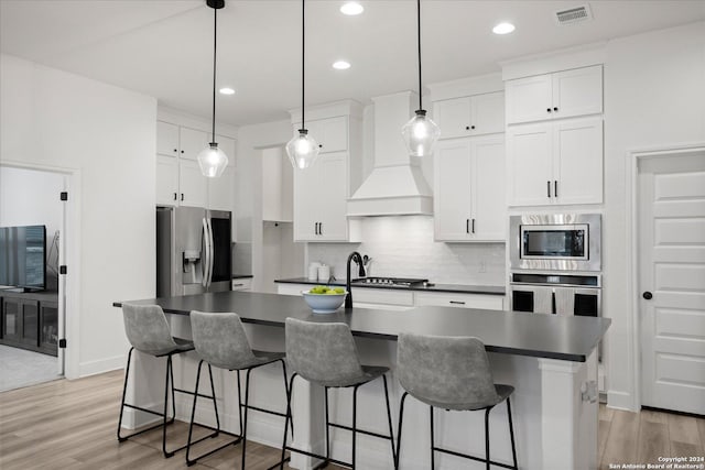 kitchen featuring appliances with stainless steel finishes, premium range hood, light hardwood / wood-style flooring, white cabinets, and an island with sink