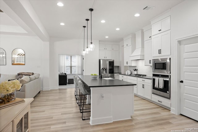 kitchen with an island with sink, decorative light fixtures, white cabinets, custom range hood, and appliances with stainless steel finishes