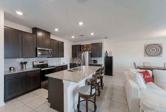 kitchen with sink, stainless steel appliances, light stone counters, an island with sink, and dark brown cabinets