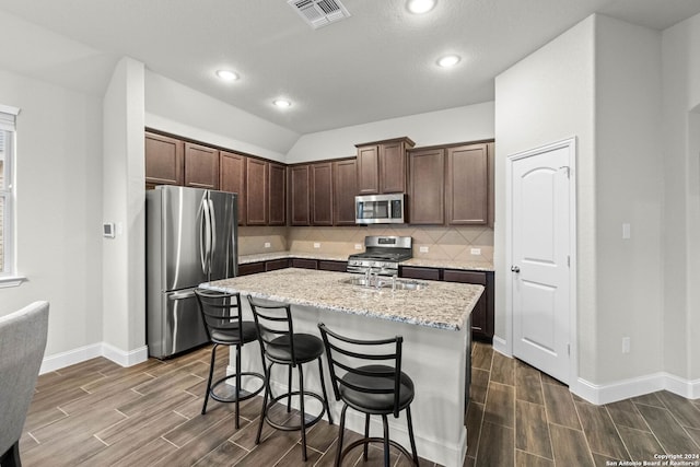 kitchen with dark hardwood / wood-style flooring, light stone counters, dark brown cabinetry, stainless steel appliances, and an island with sink