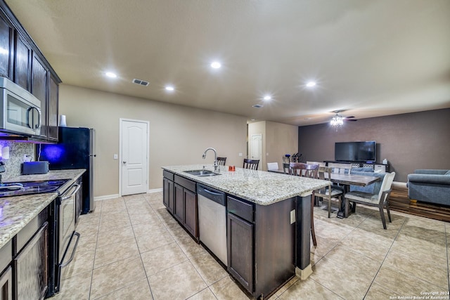 kitchen with a center island with sink, sink, dark brown cabinets, light stone counters, and stainless steel appliances