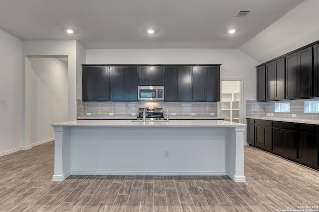 kitchen featuring tasteful backsplash, light hardwood / wood-style flooring, an island with sink, vaulted ceiling, and appliances with stainless steel finishes