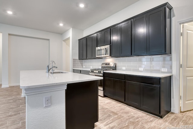 kitchen with a center island with sink, sink, decorative backsplash, light wood-type flooring, and stainless steel appliances