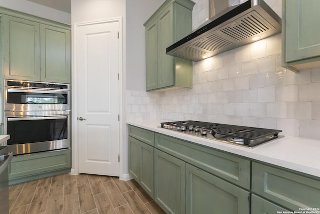 kitchen featuring stainless steel appliances, wall chimney range hood, green cabinets, backsplash, and wood-type flooring