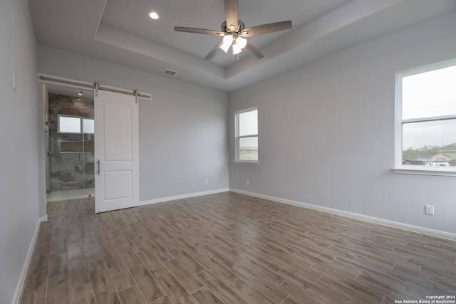 empty room featuring ceiling fan, a barn door, wood-type flooring, and a tray ceiling