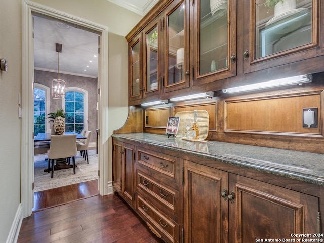 bar with crown molding, dark wood-type flooring, decorative light fixtures, dark stone countertops, and a chandelier