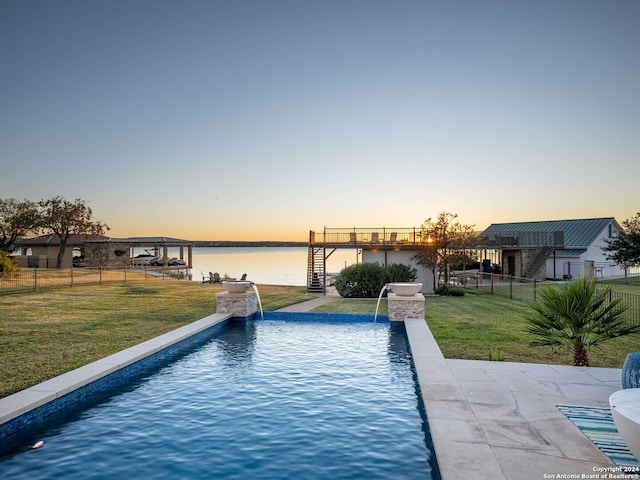 pool at dusk featuring pool water feature, a water view, a patio area, and a lawn