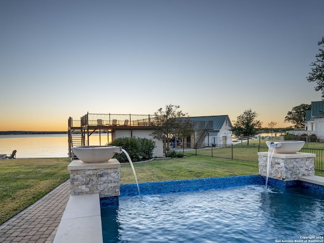 pool at dusk with pool water feature, a water view, and a yard