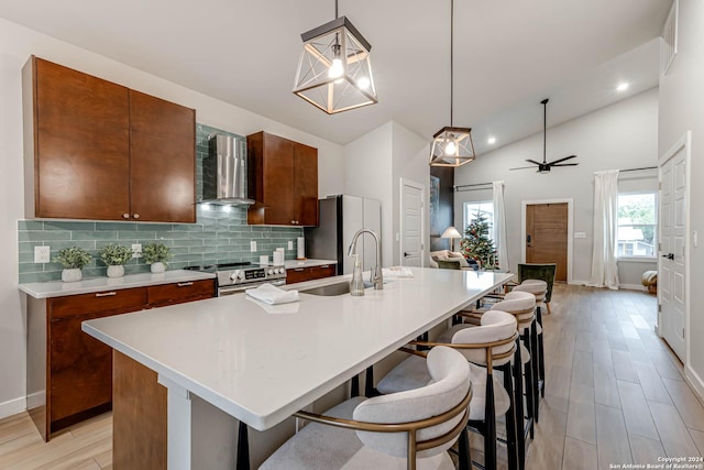 kitchen featuring wall chimney range hood, sink, hanging light fixtures, an island with sink, and appliances with stainless steel finishes