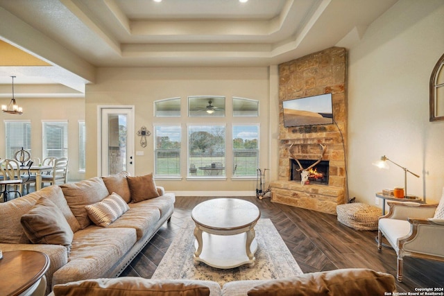 living room featuring ceiling fan with notable chandelier, a stone fireplace, a tray ceiling, and dark parquet flooring