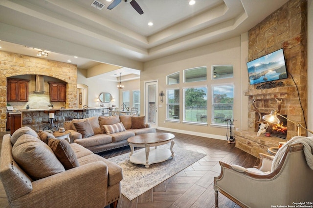 living room featuring dark parquet flooring, a raised ceiling, ceiling fan, and a stone fireplace