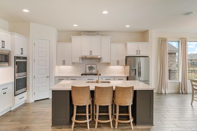 kitchen featuring tasteful backsplash, stainless steel appliances, a kitchen island with sink, white cabinets, and light hardwood / wood-style floors