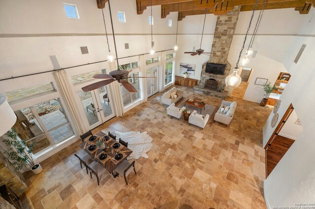 living room featuring ceiling fan, french doors, a high ceiling, a stone fireplace, and beamed ceiling