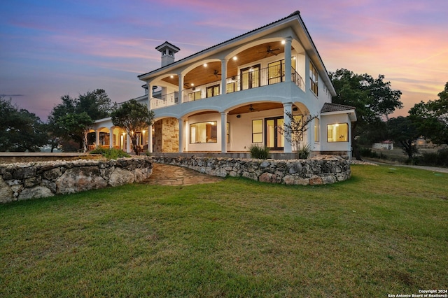 back house at dusk featuring a balcony, ceiling fan, and a lawn