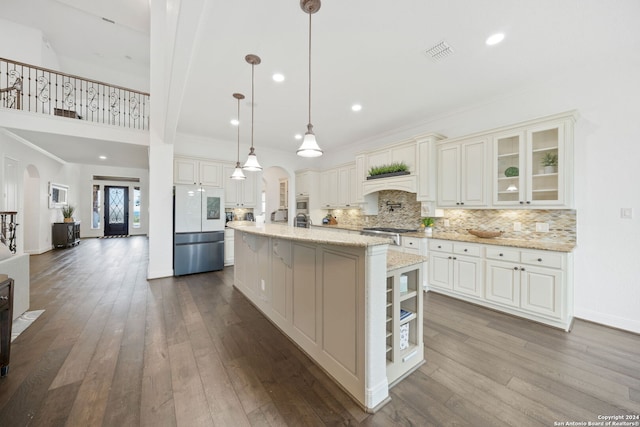 kitchen featuring stainless steel fridge, light stone counters, pendant lighting, wood-type flooring, and a center island with sink