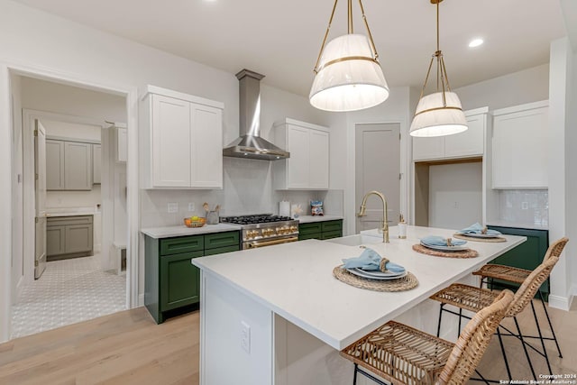 kitchen featuring high end stove, wall chimney exhaust hood, an island with sink, and decorative light fixtures