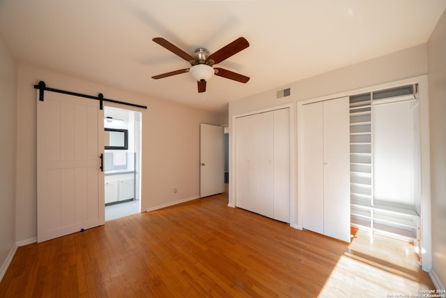 unfurnished bedroom with light wood-type flooring, ensuite bathroom, ceiling fan, a barn door, and multiple closets