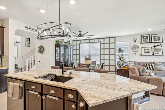 kitchen featuring pendant lighting, a kitchen island with sink, ceiling fan with notable chandelier, light stone counters, and dark brown cabinetry