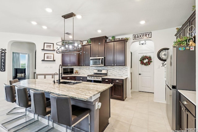 kitchen with dark brown cabinetry, stainless steel appliances, backsplash, decorative light fixtures, and a kitchen island with sink