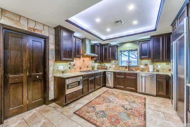 kitchen with dark brown cabinetry, wall chimney exhaust hood, backsplash, built in appliances, and a tray ceiling