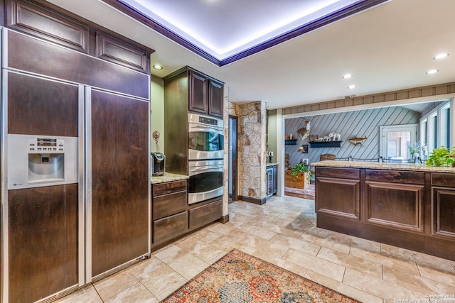 kitchen featuring paneled refrigerator, light stone counters, stainless steel double oven, dark brown cabinetry, and wood walls