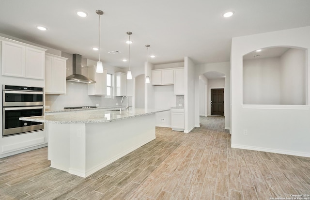 kitchen with stainless steel double oven, wall chimney range hood, an island with sink, light hardwood / wood-style floors, and white cabinets