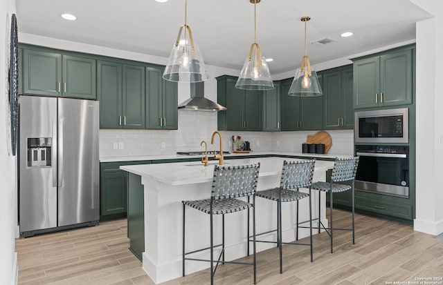 kitchen featuring a center island with sink, hanging light fixtures, wall chimney exhaust hood, appliances with stainless steel finishes, and light hardwood / wood-style floors