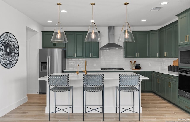 kitchen with appliances with stainless steel finishes, light wood-type flooring, wall chimney exhaust hood, sink, and an island with sink