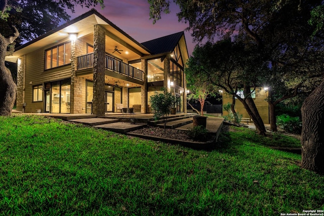 694 Scenic Cir, Pipe Creek, TX 78063 - back of property at dusk with a balcony, ceiling fan, stone siding, a yard, and a patio