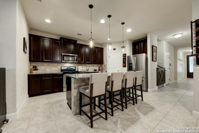 kitchen featuring pendant lighting, a kitchen island with sink, backsplash, light stone countertops, and appliances with stainless steel finishes