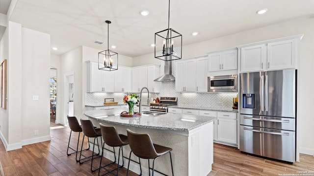 kitchen with white cabinetry, stainless steel appliances, wall chimney range hood, hardwood / wood-style floors, and a center island with sink