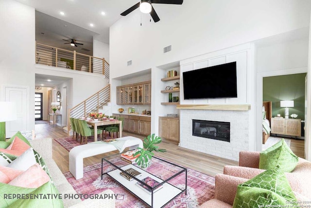 living room with a towering ceiling, light hardwood / wood-style floors, a brick fireplace, and ceiling fan