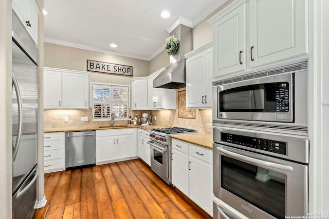 kitchen with built in appliances, white cabinetry, sink, and wall chimney range hood