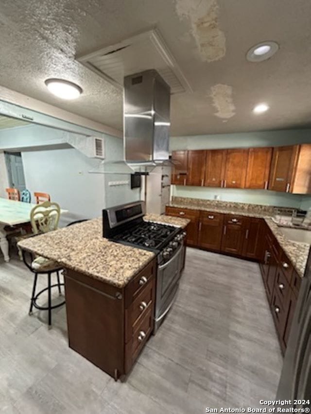 kitchen with wall chimney exhaust hood, light stone countertops, a textured ceiling, and stainless steel gas range