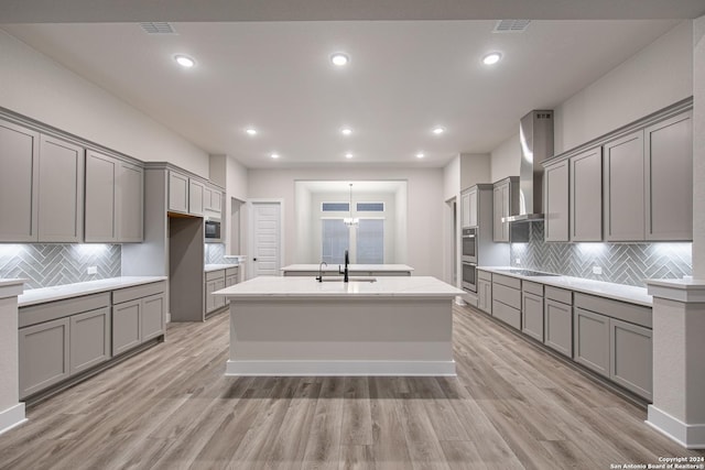 kitchen with gray cabinetry, a center island with sink, wall chimney range hood, sink, and light hardwood / wood-style floors