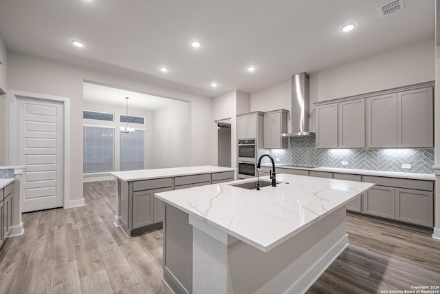 kitchen featuring light stone counters, gray cabinetry, wall chimney exhaust hood, light hardwood / wood-style floors, and an island with sink