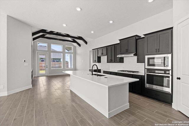 kitchen featuring sink, stainless steel appliances, beamed ceiling, a kitchen island with sink, and light wood-type flooring