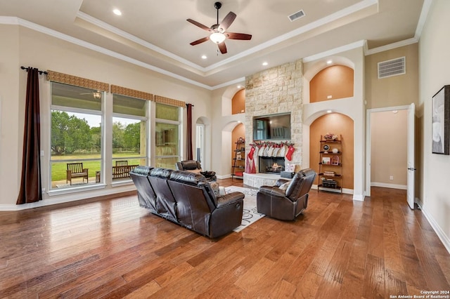 living room with a stone fireplace, crown molding, hardwood / wood-style flooring, ceiling fan, and a tray ceiling