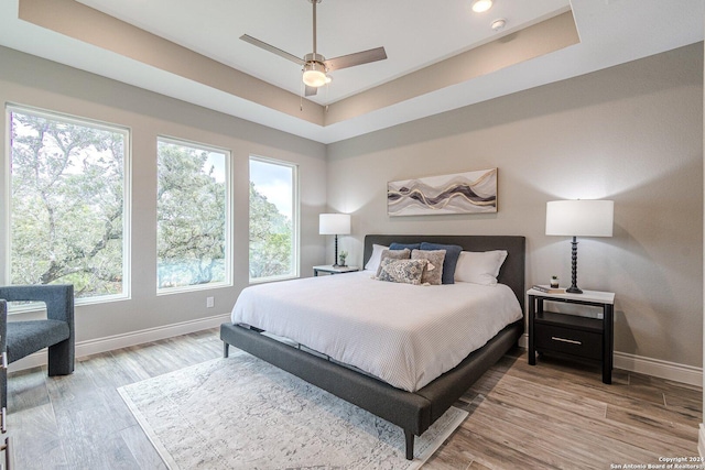 bedroom with ceiling fan, a raised ceiling, light hardwood / wood-style flooring, and multiple windows