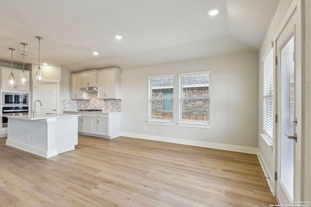 kitchen featuring built in microwave, stainless steel oven, hanging light fixtures, an island with sink, and white cabinets